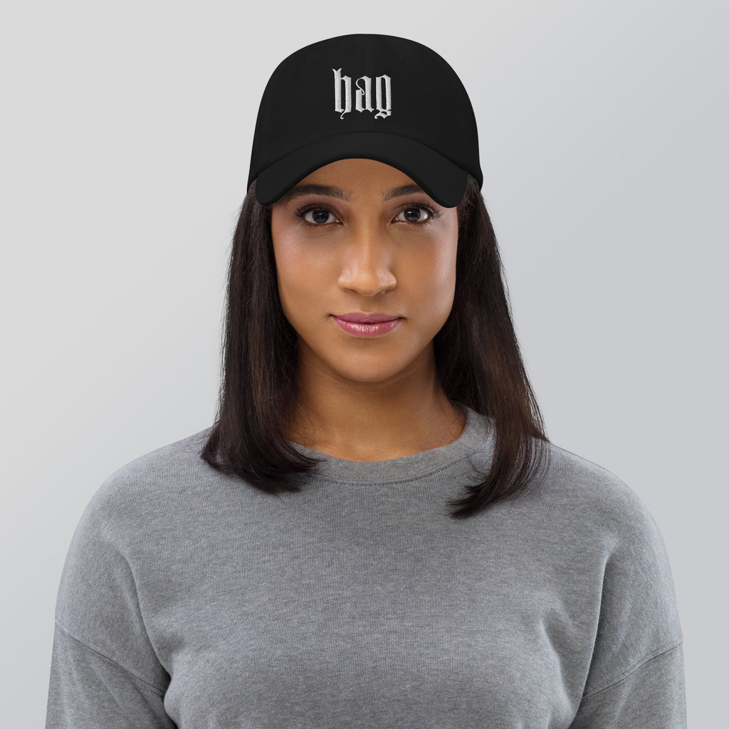 Black dad hat embroidered with the word ‘HAG’ in ornate vintage gothic lettering. Alternative fashion accessory with queer streetwear aesthetic, witchy fashion, sarcastic humor, feminist style, indie fashion, spooky cute apparel, gothic embroidery, punk inspired streetwear, adjustable baseball cap, casual everyday alternative outfit, internet humor fashion, artsy accessory, and edgy statement hat for concerts, festivals, casual wear, and alternative wardrobes.