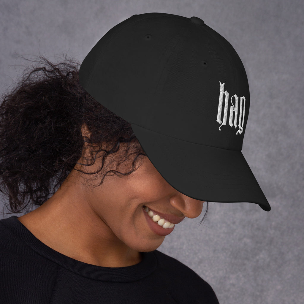 Black dad hat embroidered with the word ‘HAG’ in ornate vintage gothic lettering. Alternative fashion accessory with queer streetwear aesthetic, witchy fashion, sarcastic humor, feminist style, indie fashion, spooky cute apparel, gothic embroidery, punk inspired streetwear, adjustable baseball cap, casual everyday alternative outfit, internet humor fashion, artsy accessory, and edgy statement hat for concerts, festivals, casual wear, and alternative wardrobes.