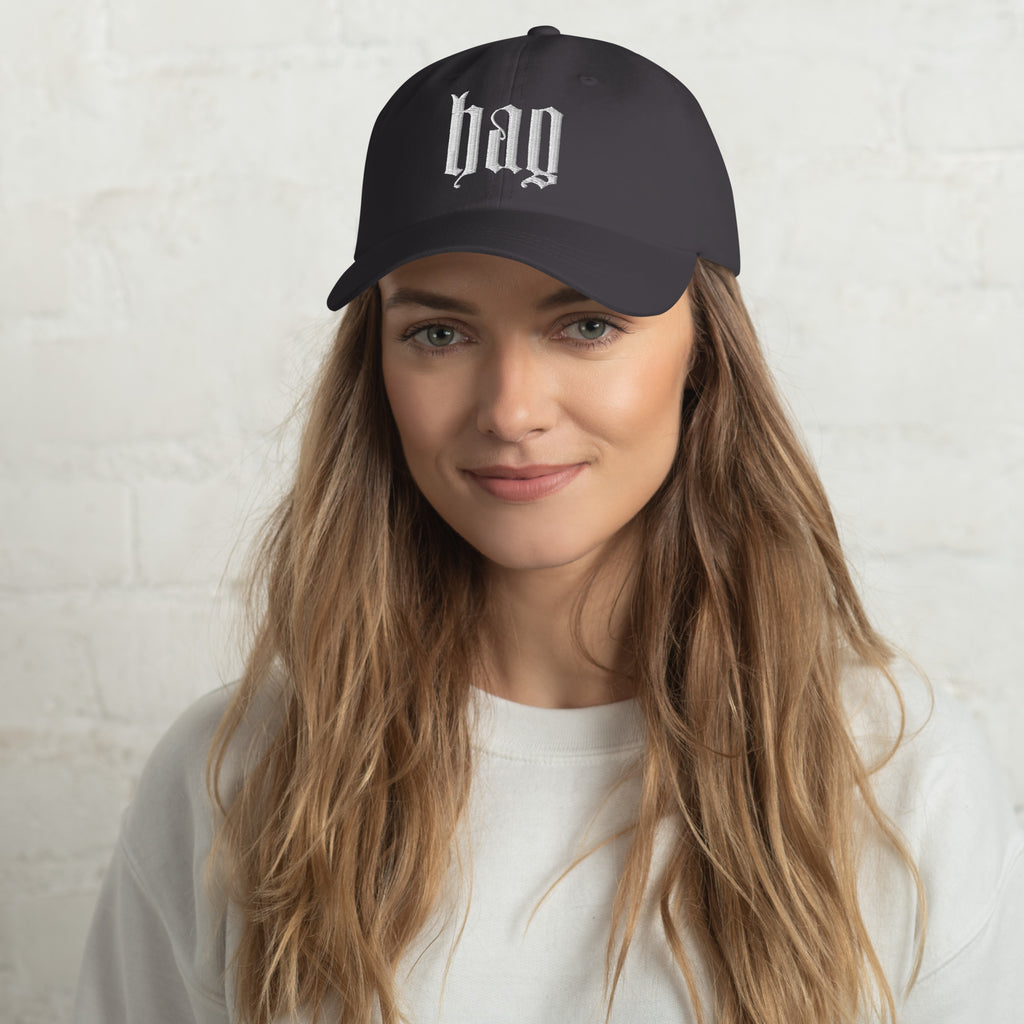 Black dad hat embroidered with the word ‘HAG’ in ornate vintage gothic lettering. Alternative fashion accessory with queer streetwear aesthetic, witchy fashion, sarcastic humor, feminist style, indie fashion, spooky cute apparel, gothic embroidery, punk inspired streetwear, adjustable baseball cap, casual everyday alternative outfit, internet humor fashion, artsy accessory, and edgy statement hat for concerts, festivals, casual wear, and alternative wardrobes.
