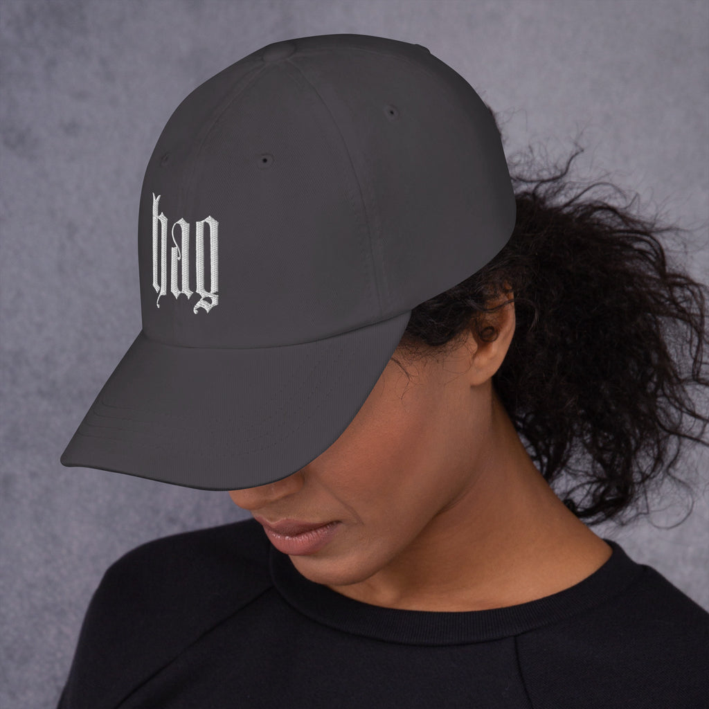 Black dad hat embroidered with the word ‘HAG’ in ornate vintage gothic lettering. Alternative fashion accessory with queer streetwear aesthetic, witchy fashion, sarcastic humor, feminist style, indie fashion, spooky cute apparel, gothic embroidery, punk inspired streetwear, adjustable baseball cap, casual everyday alternative outfit, internet humor fashion, artsy accessory, and edgy statement hat for concerts, festivals, casual wear, and alternative wardrobes.