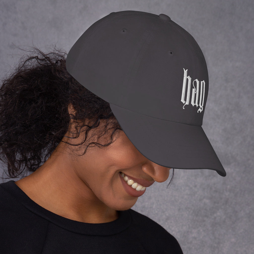 Black dad hat embroidered with the word ‘HAG’ in ornate vintage gothic lettering. Alternative fashion accessory with queer streetwear aesthetic, witchy fashion, sarcastic humor, feminist style, indie fashion, spooky cute apparel, gothic embroidery, punk inspired streetwear, adjustable baseball cap, casual everyday alternative outfit, internet humor fashion, artsy accessory, and edgy statement hat for concerts, festivals, casual wear, and alternative wardrobes.