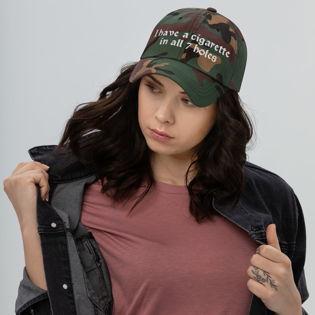 Baseball cap embroidered with the phrase ‘I Have a Cigarette in All 7 Holes’ in ornate vintage gothic lettering. Alternative fashion accessory with edgy internet humor, queer streetwear aesthetic, punk inspired style, ironic meme fashion, tattoo inspired typography, goth streetwear, adjustable dad hat, casual alternative outfit accessory, artsy statement cap, chaotic humor apparel, indie fashion, and expressive alternative clothing for concerts, festivals, everyday wear, and alternative wardrobes.
