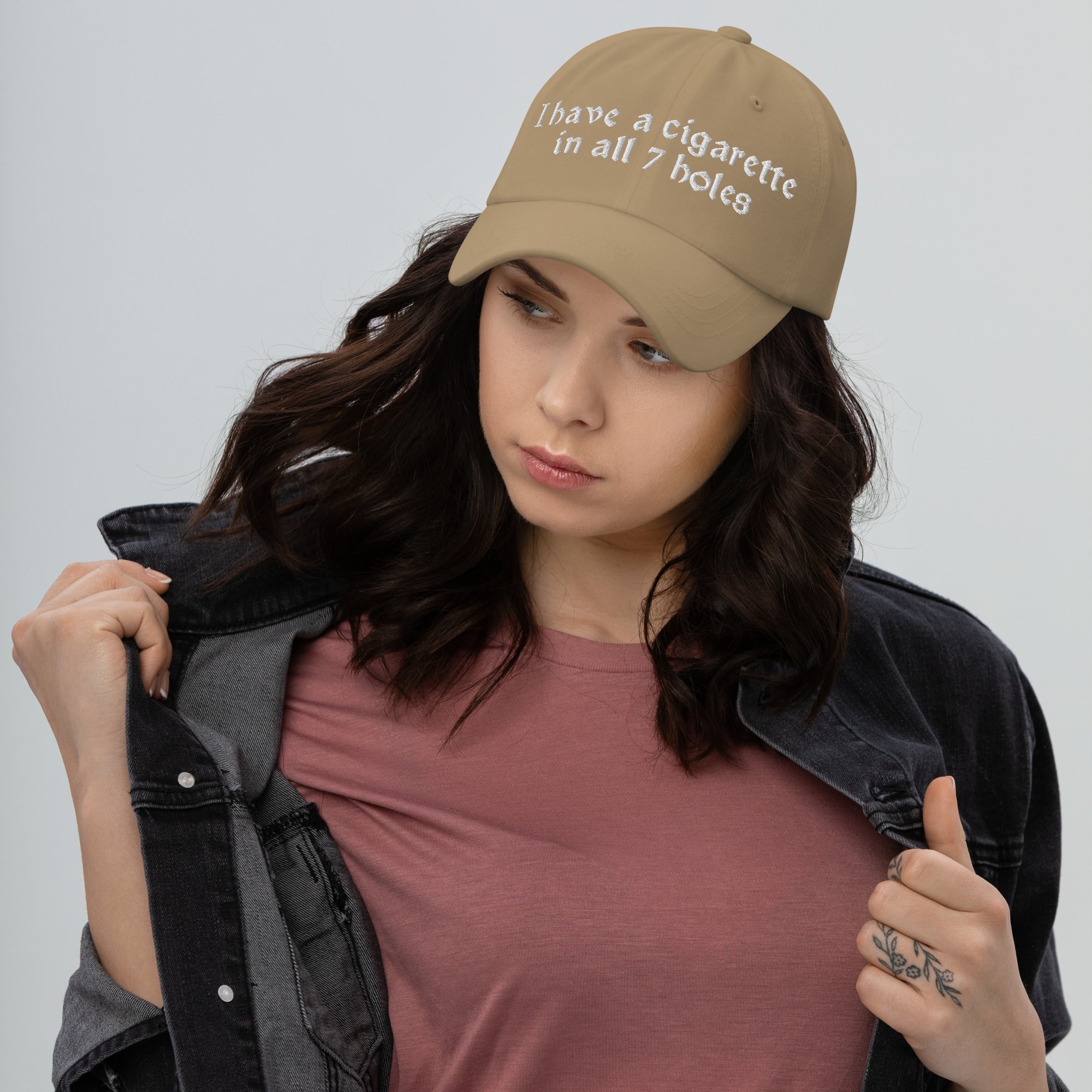 Baseball cap embroidered with the phrase ‘I Have a Cigarette in All 7 Holes’ in ornate vintage gothic lettering. Alternative fashion accessory with edgy internet humor, queer streetwear aesthetic, punk inspired style, ironic meme fashion, tattoo inspired typography, goth streetwear, adjustable dad hat, casual alternative outfit accessory, artsy statement cap, chaotic humor apparel, indie fashion, and expressive alternative clothing for concerts, festivals, everyday wear, and alternative wardrobes.