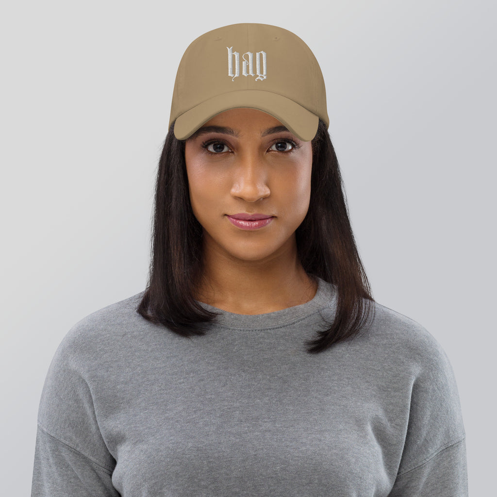 Black dad hat embroidered with the word ‘HAG’ in ornate vintage gothic lettering. Alternative fashion accessory with queer streetwear aesthetic, witchy fashion, sarcastic humor, feminist style, indie fashion, spooky cute apparel, gothic embroidery, punk inspired streetwear, adjustable baseball cap, casual everyday alternative outfit, internet humor fashion, artsy accessory, and edgy statement hat for concerts, festivals, casual wear, and alternative wardrobes.