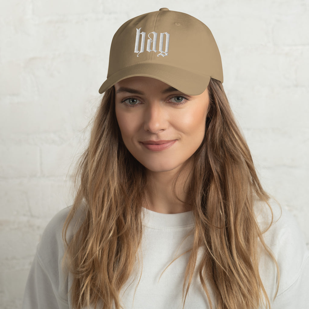 Black dad hat embroidered with the word ‘HAG’ in ornate vintage gothic lettering. Alternative fashion accessory with queer streetwear aesthetic, witchy fashion, sarcastic humor, feminist style, indie fashion, spooky cute apparel, gothic embroidery, punk inspired streetwear, adjustable baseball cap, casual everyday alternative outfit, internet humor fashion, artsy accessory, and edgy statement hat for concerts, festivals, casual wear, and alternative wardrobes.