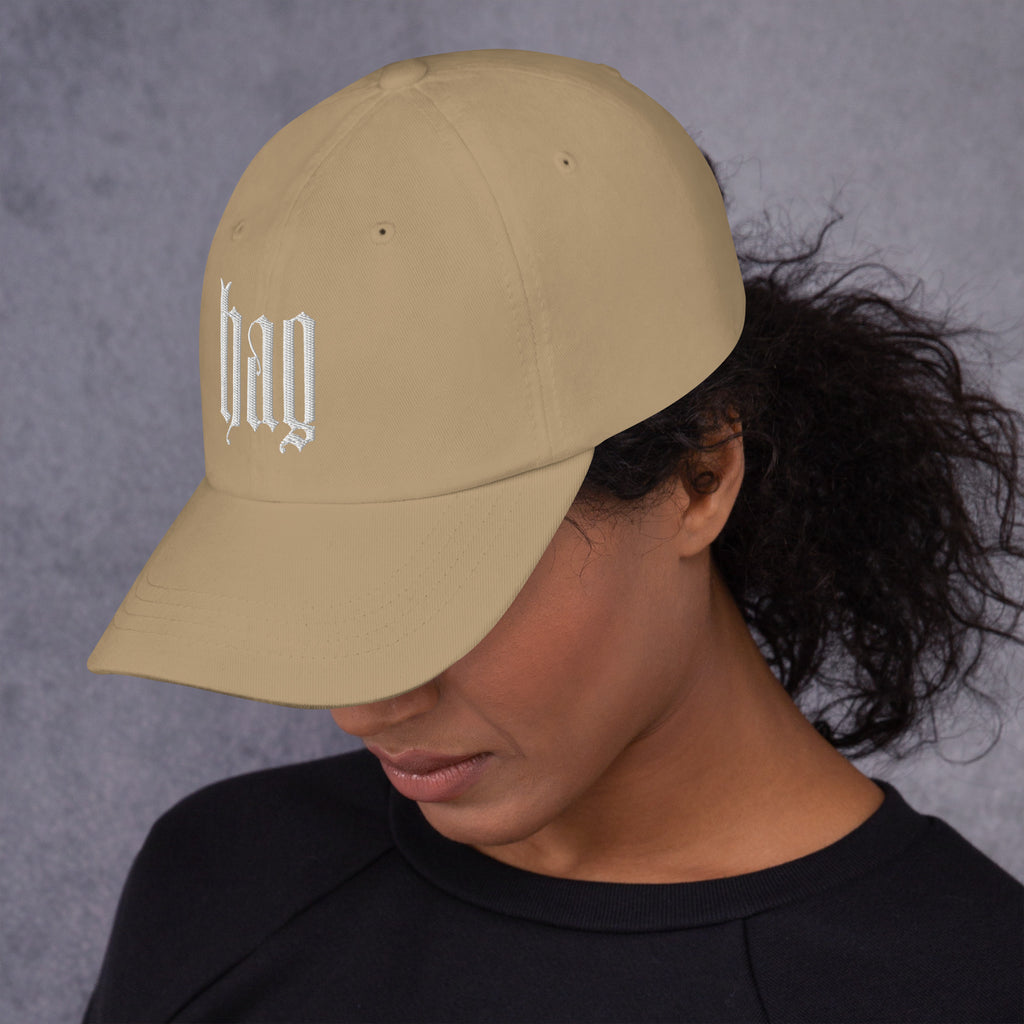 Black dad hat embroidered with the word ‘HAG’ in ornate vintage gothic lettering. Alternative fashion accessory with queer streetwear aesthetic, witchy fashion, sarcastic humor, feminist style, indie fashion, spooky cute apparel, gothic embroidery, punk inspired streetwear, adjustable baseball cap, casual everyday alternative outfit, internet humor fashion, artsy accessory, and edgy statement hat for concerts, festivals, casual wear, and alternative wardrobes.