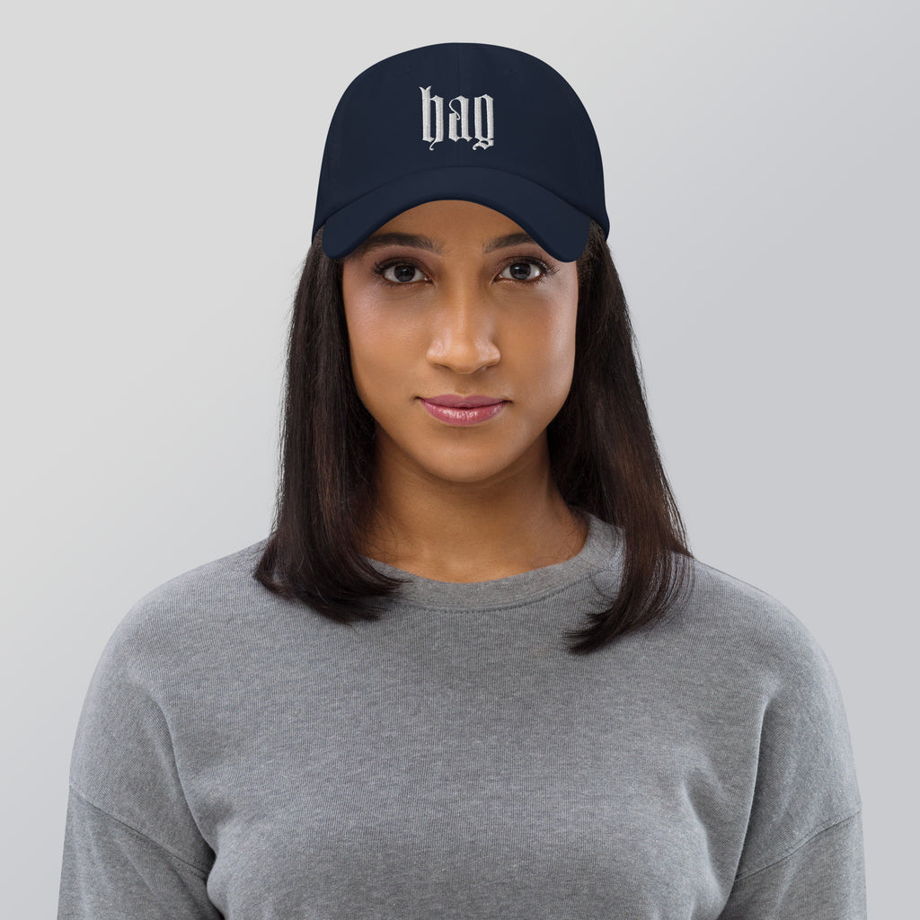 Black dad hat embroidered with the word ‘HAG’ in ornate vintage gothic lettering. Alternative fashion accessory with queer streetwear aesthetic, witchy fashion, sarcastic humor, feminist style, indie fashion, spooky cute apparel, gothic embroidery, punk inspired streetwear, adjustable baseball cap, casual everyday alternative outfit, internet humor fashion, artsy accessory, and edgy statement hat for concerts, festivals, casual wear, and alternative wardrobes.
