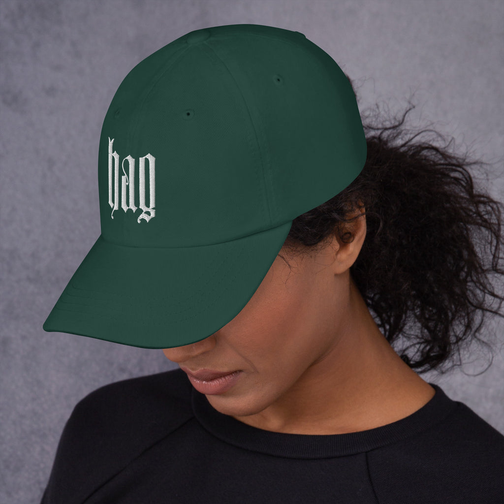 Black dad hat embroidered with the word ‘HAG’ in ornate vintage gothic lettering. Alternative fashion accessory with queer streetwear aesthetic, witchy fashion, sarcastic humor, feminist style, indie fashion, spooky cute apparel, gothic embroidery, punk inspired streetwear, adjustable baseball cap, casual everyday alternative outfit, internet humor fashion, artsy accessory, and edgy statement hat for concerts, festivals, casual wear, and alternative wardrobes.