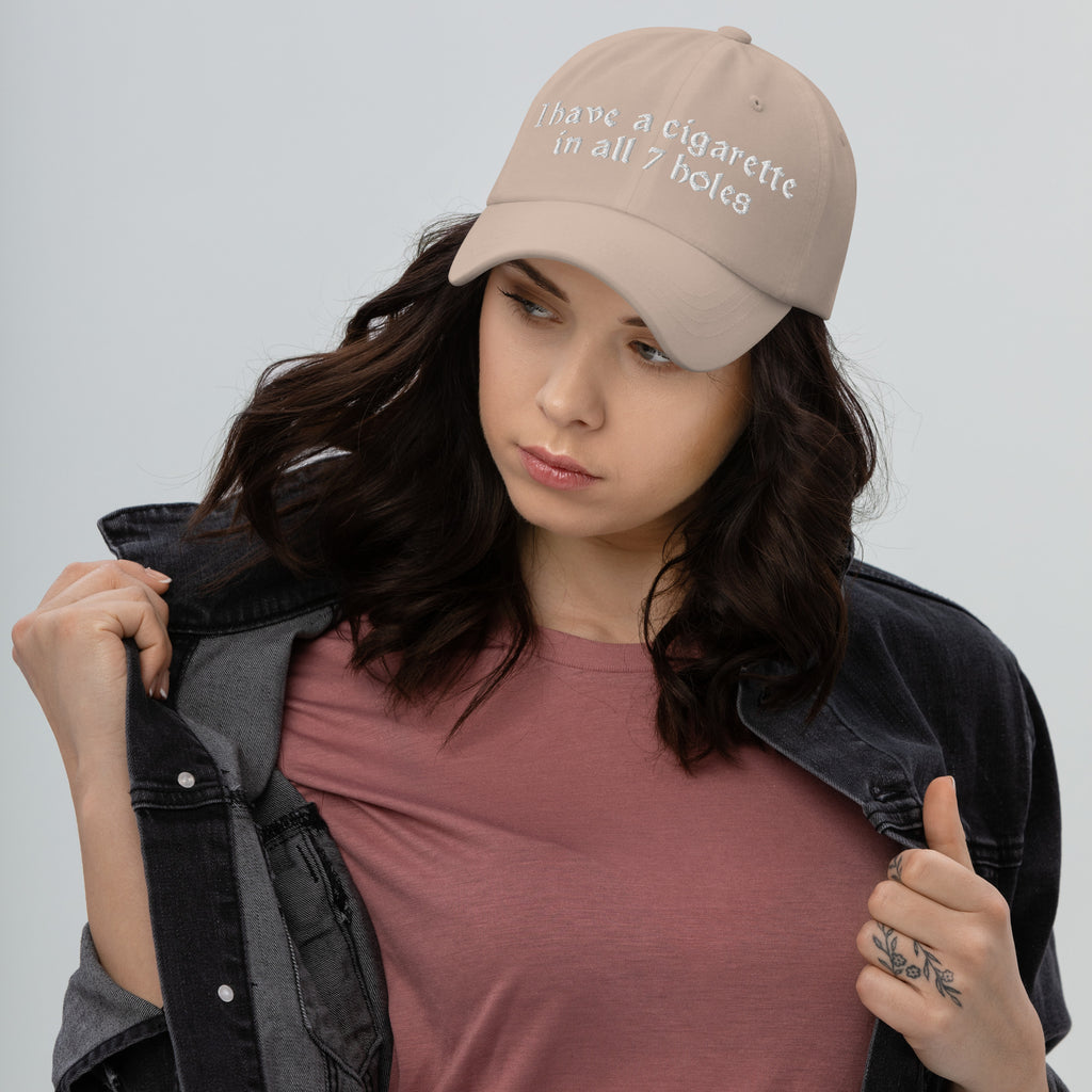 Baseball cap embroidered with the phrase ‘I Have a Cigarette in All 7 Holes’ in ornate vintage gothic lettering. Alternative fashion accessory with edgy internet humor, queer streetwear aesthetic, punk inspired style, ironic meme fashion, tattoo inspired typography, goth streetwear, adjustable dad hat, casual alternative outfit accessory, artsy statement cap, chaotic humor apparel, indie fashion, and expressive alternative clothing for concerts, festivals, everyday wear, and alternative wardrobes.
